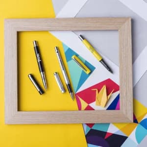 A collection of pens in black, silver, and yellow arranged within a wooden frame on a yellow geometric background.