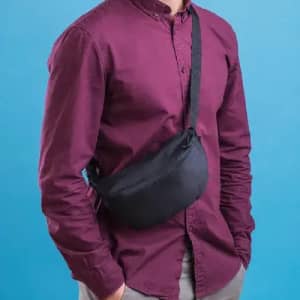 A sling bag in black worn across the body by a person in a maroon shirt, against a blue background.