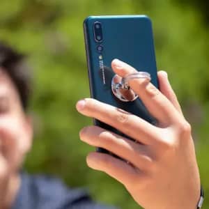 A smartphone with a blue back, featuring a ring grip on the rear, held by a person against a green background.