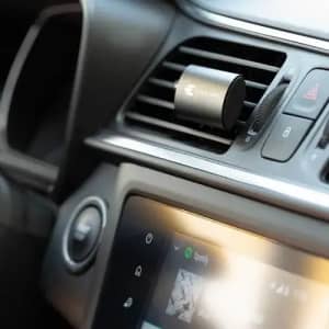 A car air vent with a black circular accessory attached, next to a touchscreen displaying a music app.