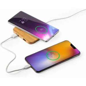 A wireless charging pad with two smartphones charging simultaneously. The pad is wooden, and the phones display battery percentages.