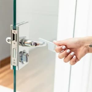 A hand holds a white door lock tool, with a shiny chrome door handle and glass door visible in the image.