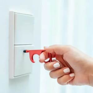 A red touchless tool is being used to press a light switch. The tool features a hook and a keyring attachment.