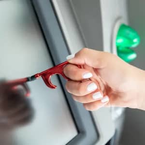 A red ATM tool is held by a hand, designed to assist with touchscreens and reduce contact.
