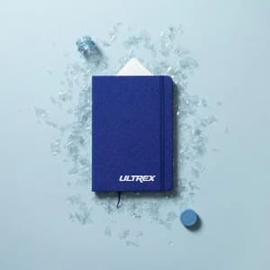 A navy blue notebook is placed on a light blue surface surrounded by shards of glass and a small blue bottle.