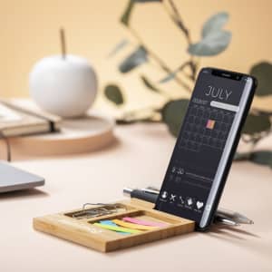 A bamboo organiser featuring sticky notes, a pen, a paper clip, and a mobile phone displaying a calendar.
