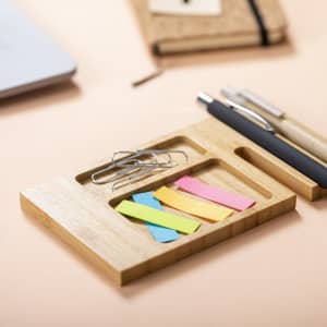 A bamboo organiser with paper clips, colourful sticky notes, a pen, and a notebook on a light background.