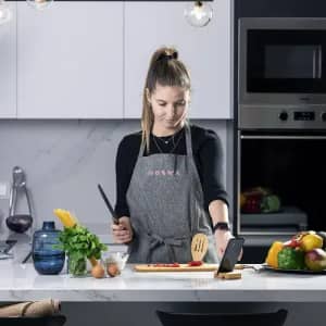 A woman in an apron stands at a kitchen counter, chopping vegetables with a knife, surrounded by fresh ingredients and cookware.