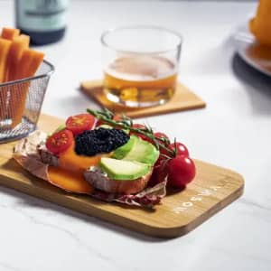 A wooden serving board with a slice of bread topped with avocado, tomatoes, caviar, and sauce, alongside a drink.