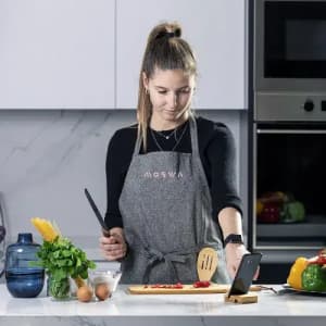 A woman in a grey apron prepares vegetables on a wooden board in a modern kitchen. A phone is positioned nearby.