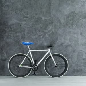 A bicycle with a white frame, black wheels, and a blue seat, positioned against a textured grey wall.
