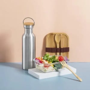 A drink bottle in stainless steel with a wooden lid, accompanied by a salad bowl and bamboo utensils on a pastel background.