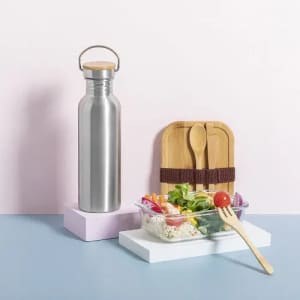 A drink bottle in stainless steel with a wooden lid, beside a glass container of fresh salad on a pastel background.