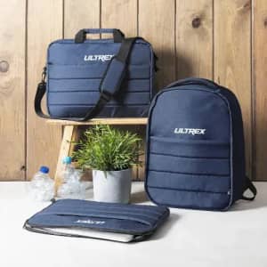 A navy blue backpack, bag, and laptop sleeve are displayed on a wooden stand with a small plant and water bottles.