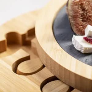 A wooden cheese board featuring a round design, with sliced cheese and a fig on a dark slate surface.