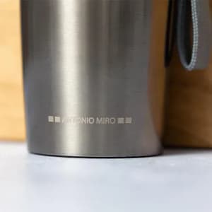 A drink bottle in stainless steel with a smooth finish and a grey strap, featuring a logo at the base.