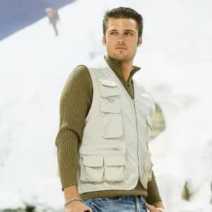 A man wearing a beige vest over a green sweater stands against a snowy backdrop. The vest has multiple pockets and a logo.