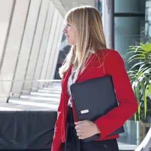 A woman in a red blazer holds a black folder, standing in a modern building with large windows and greenery.