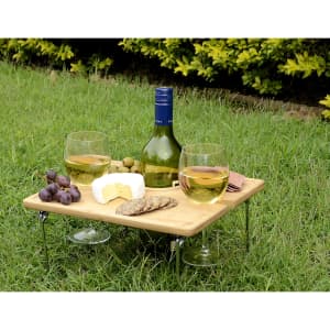 A wooden picnic table with two glasses of white wine, grapes, olives, cheese, and crackers on a grassy surface.