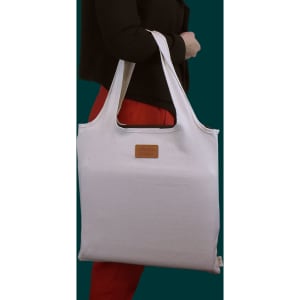 A white tote bag with long handles, featuring a logo, held by a person wearing a black top and red pants.