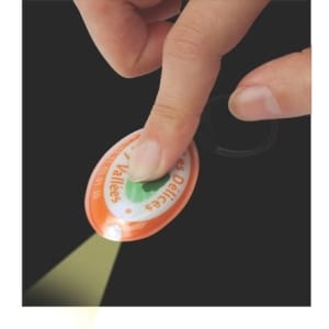 A hand pressing an orange press-light keyring, emitting light. The keyring features a logo on its surface.