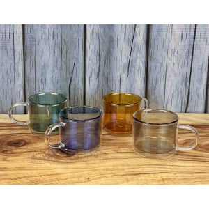 Four transparent double-wall mugs in green, amber, blue, and grey arranged on a wooden surface against a textured background.