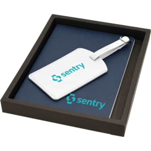 A navy notebook and white luggage tag with a branding logo, presented in a dark box.