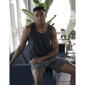 A black unisex tank top is worn by a model sitting on a blue couch, surrounded by indoor plants.