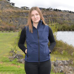 A navy blue softshell vest with a zip and pockets, worn by a model in an outdoor setting near water and greenery.