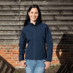 A navy blue ladies softshell jacket with a zip front and a logo, worn by a woman standing against a wooden background.
