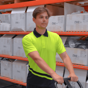 A man in a high-visibility yellow and navy polo shirt stands next to a trolley in a warehouse. The shirt has a logo.