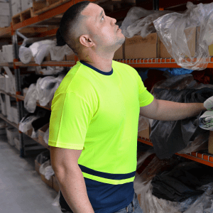 A man wearing a bright yellow and navy work t-shirt stands in a storage area, reaching for items on a shelf.