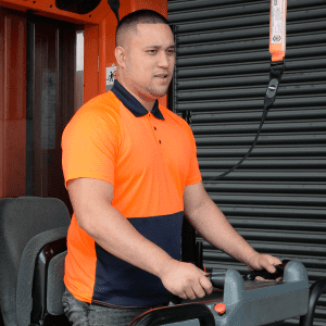 A man wearing an orange and navy blue polo shirt operates a forklift in a work environment. The shirt has a logo.