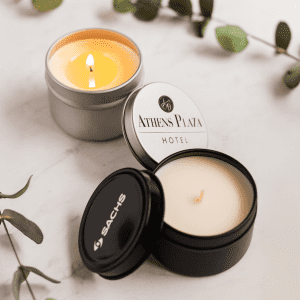 Three scented candles in silver and black containers, one with a lit wick, accompanied by green leaves.
