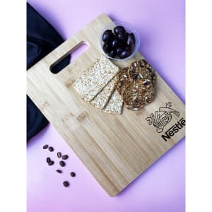 A bamboo chopping board featuring a logo, with crackers, seed biscuits, and a bowl of olives on a pink surface.
