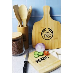 A bamboo cutting board with a handle, surrounded by kitchen utensils, sliced vegetables, and a glass container.