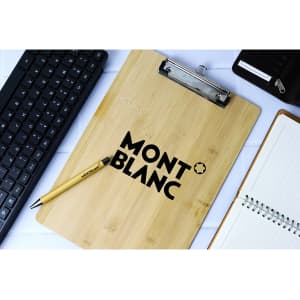 A bamboo clipboard with a metal clip and a logo, alongside a keyboard, pen, and notepad.