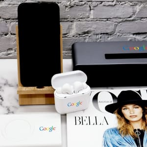 A bamboo phone stand with a smartphone, white earbuds case, and a magazine on a marble surface, featuring logos.