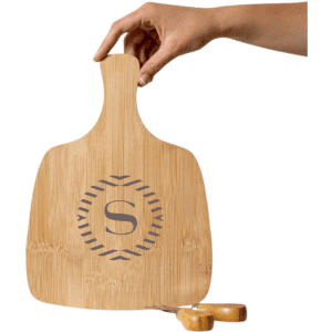 A bamboo cheeseboard with an engraved logo and a hand holding it. Two wooden cheese knives are nearby.