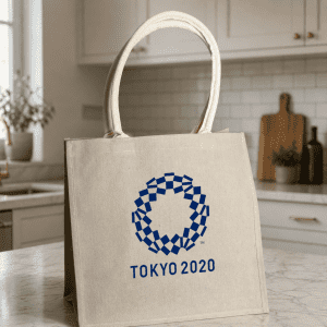 A jute tote bag in a natural colour with a logo design in blue. Features sturdy handles and a structured shape.