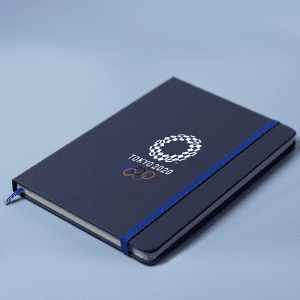 A navy A5 notebook with an elastic band closure and an Olympic-themed logo on the cover.