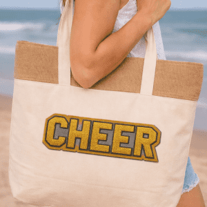 A jute and cotton cooler bag in natural colour, featuring a gold and grey logo, held by a person at the beach.