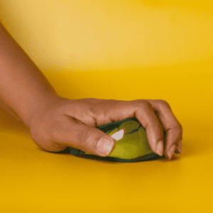 A green jelly cube sensory toy being squeezed by a hand, set against a bright yellow background.