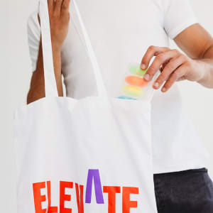 A white tote bag with colourful lettering, being held by a person as they retrieve a clear container with coloured items.