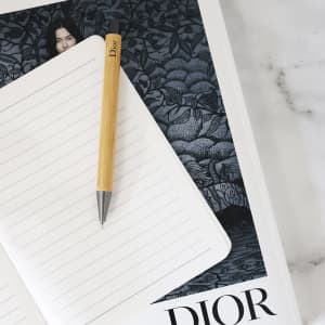 A wooden bamboo pen with a silver tip rests on an open lined notebook against a decorative background. It features a logo.