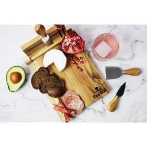 A wooden cheeseboard with cheese, meats, avocado, bread, and a pomegranate, featuring utensils and a logo.