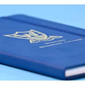 A blue A5 notebook with a logo and gold lettering on the cover, featuring a textured finish.