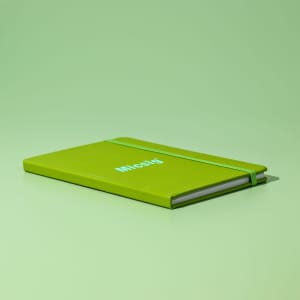 A green A5 notebook with an elastic band and a simple logo on the cover, resting on a matching green surface.