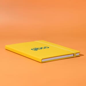 A yellow A5 notebook with an elastic band closure and a logo on the front, set against an orange background.