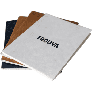 A set of three A5 notebooks in white, black, and brown, featuring a logo, placed on a desk with a coffee cup.